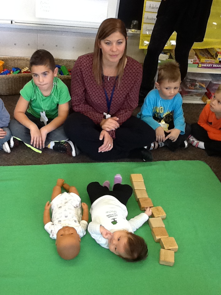 A woman sits with two students while two babies lie on a math