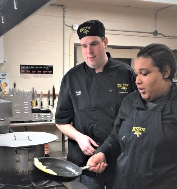 A teacher in a culinary class works with a student at a stove 